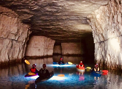 A group kayaks in an underground pool with individual kayaks, headlamps, and other caving gear.