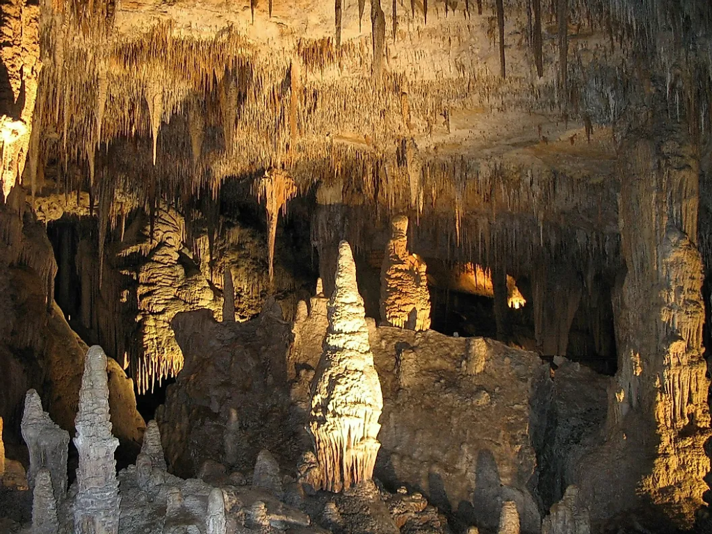 An image shows several thick stalagmites with a wide variety of thinner stalactites hanging from the ceiling above inside a cave.
